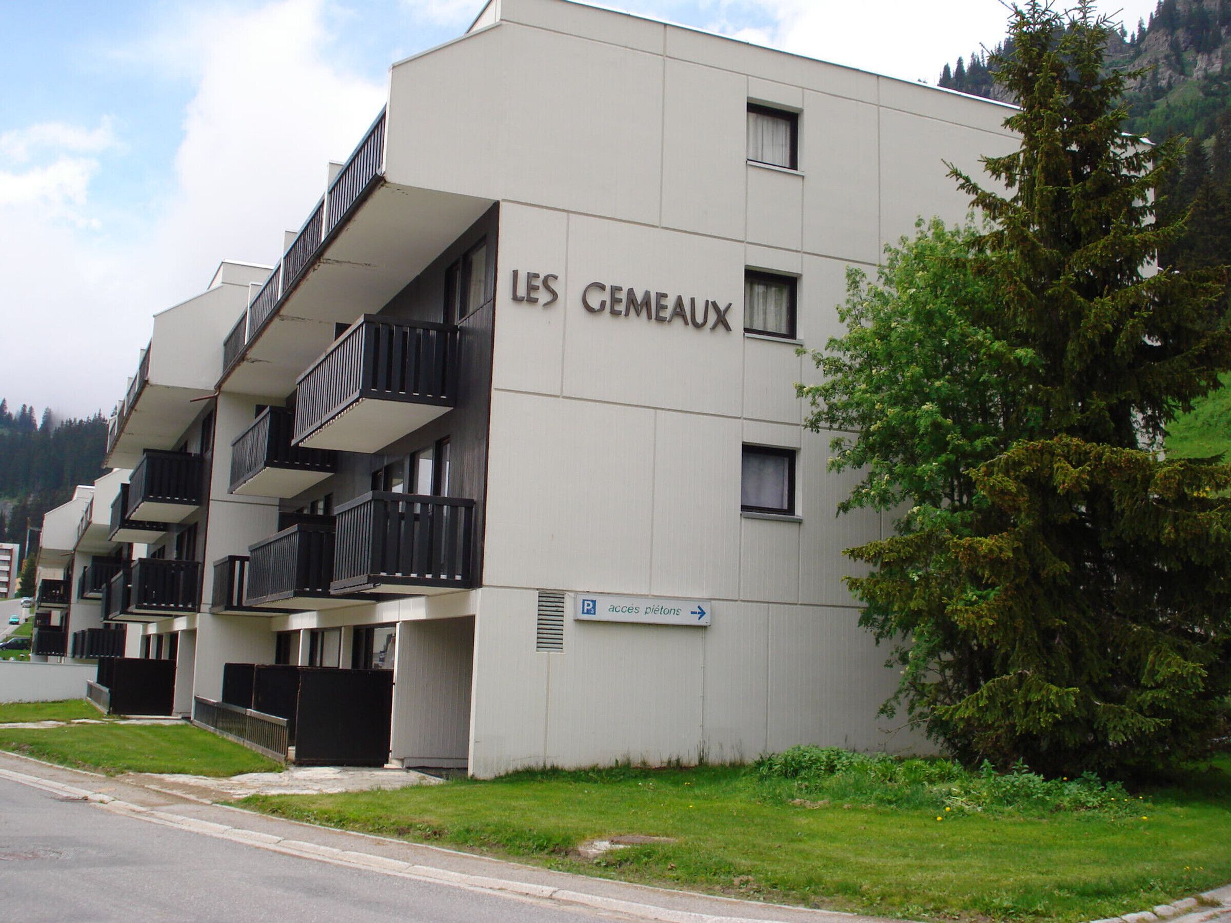 Un edificio moderno llamado "LES CEMEAUX" con varios balcones y un letrero que indica acceso a una piscina.