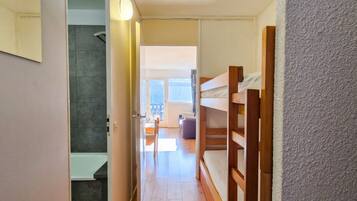 Un intérieur moderne avec un lit superposé en bois, une salle de bain avec douche et une vue sur un espace de vie comprenant un canapé et une table.