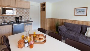 Una cocina acogedora con armarios de madera, un microondas y una cafetera. Hay una mesa de comedor con opciones para el desayuno, como cruasanes y jugo. Un sofá gris está apoyado contra la pared, y una fotografía enmarcada cuelga sobre él.