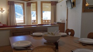 Ein Essbereich mit einem Holztisch, gedeckt für vier Personen, einem an der Wand befestigten Fernseher und einem Fenster mit Blick auf schneebedeckte Berge.