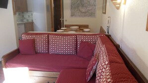 Ein gemütliches Wohnzimmer mit einem roten, gemusterten Sofa, einem Holzesstisch mit Tellern und einer Schüssel sowie einer Karte an der Wand.