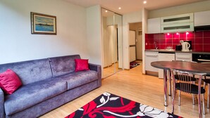 A modern living room with a grey sofa and red cushions, a wooden floor, and a kitchen with red tiles and stainless steel appliances. - Renovated studio for 4, near slopes in Flaine (Flaine)