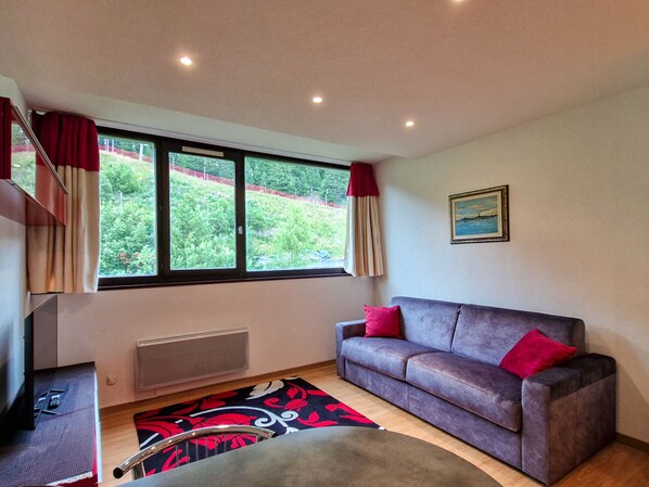 A room with a grey sofa, red cushions, a window with curtains, a radiator, and a painting on the wall.