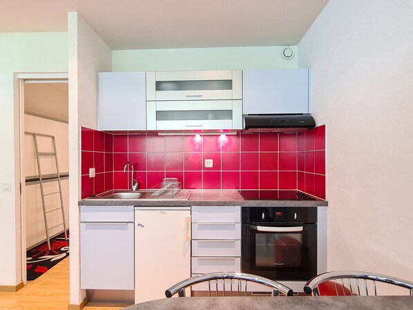 A compact kitchen with white cabinets, a red-tiled backsplash, a stainless steel sink, a dishwasher, and a built-in oven.