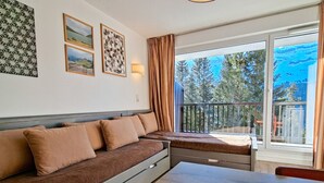 Habitación moderna con sofá, una mesa de comedor y balcón con vistas a árboles y montañas.
