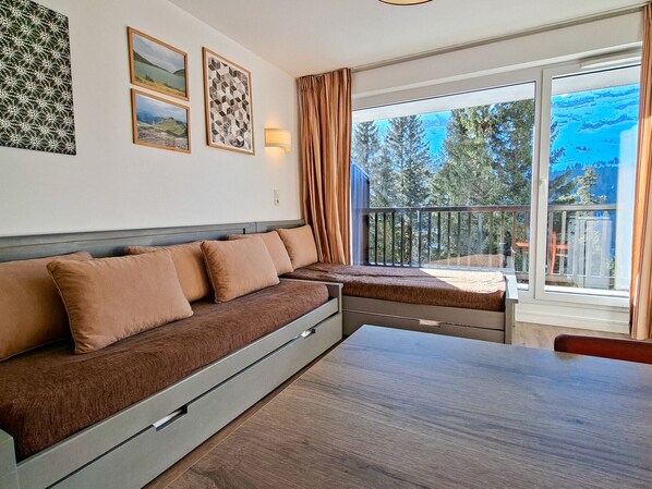 A modern room with a sofa, a dining table, and a balcony with a view of trees and mountains.