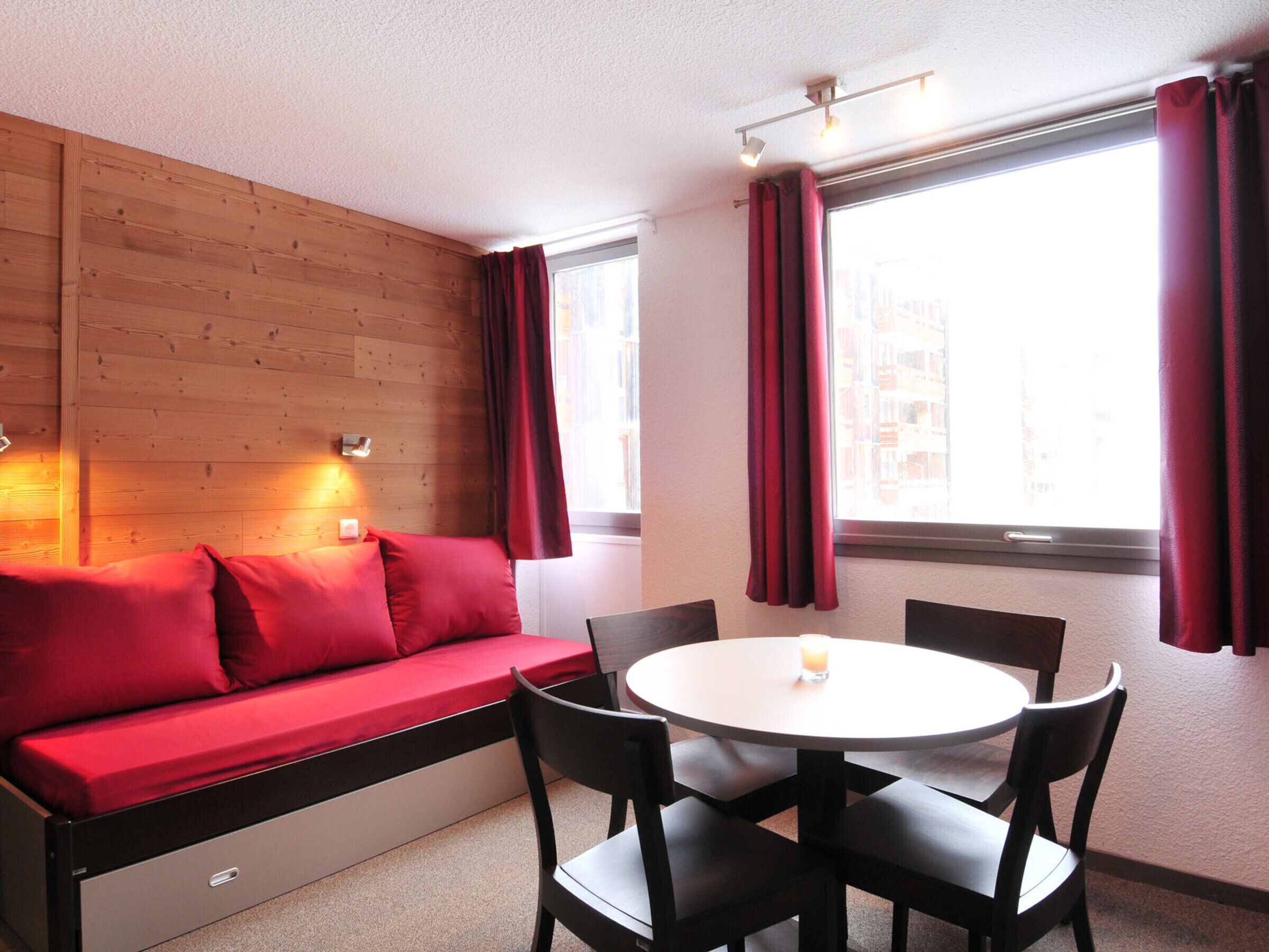 Une pièce avec un mur en bois, un canapé rouge, une table ronde entourée de chaises et une fenêtre avec des rideaux.