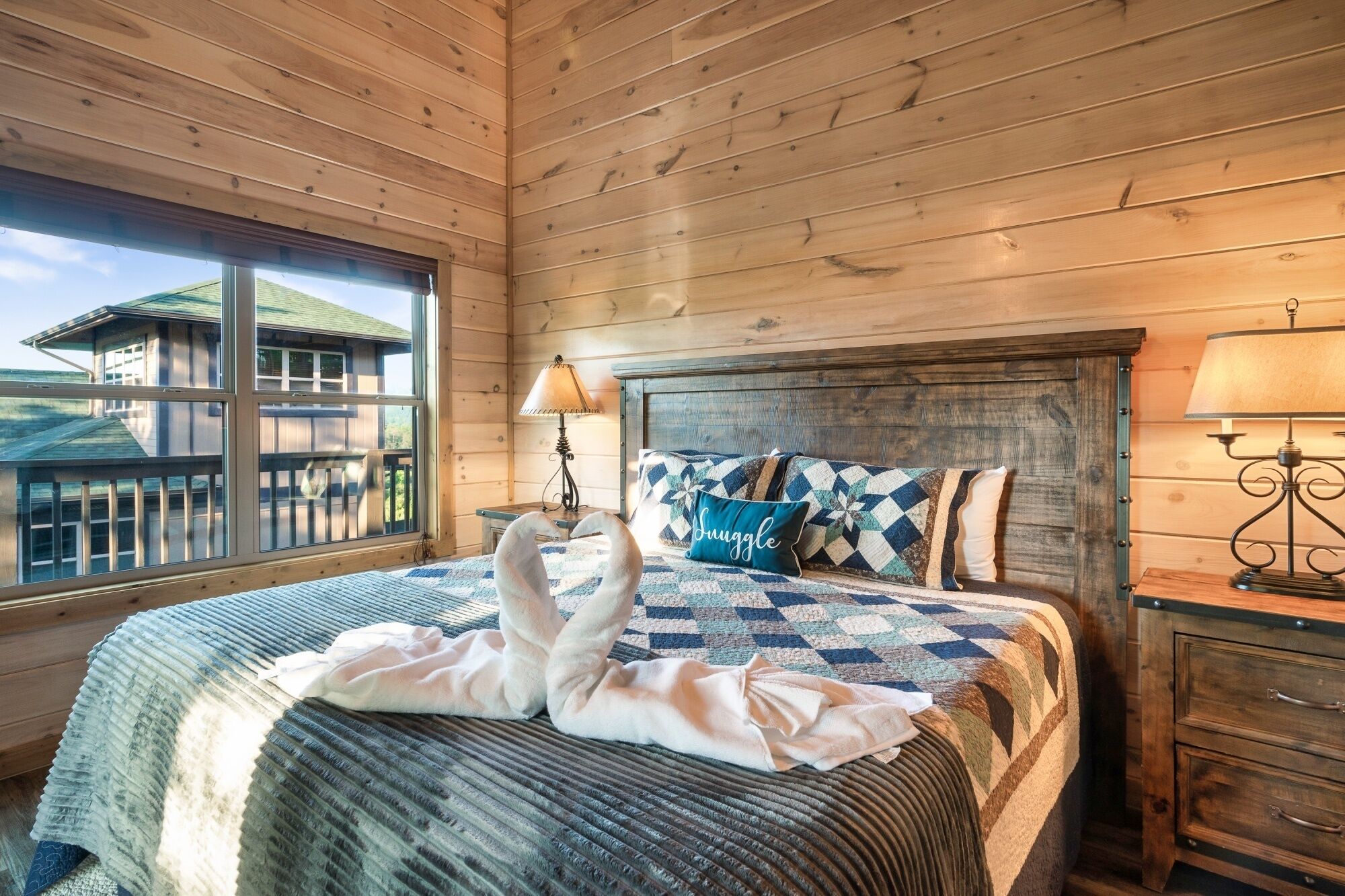 A wooden cabin-style bedroom with a large bed, two bedside lamps, and a view of a wooden deck outside.