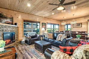 A cozy log cabin interior with a stone fireplace, leather furniture, and large windows offering a view of the outdoors. - Hummingbird Hill Vista- Pool HotTub View (Pigeon Forge)