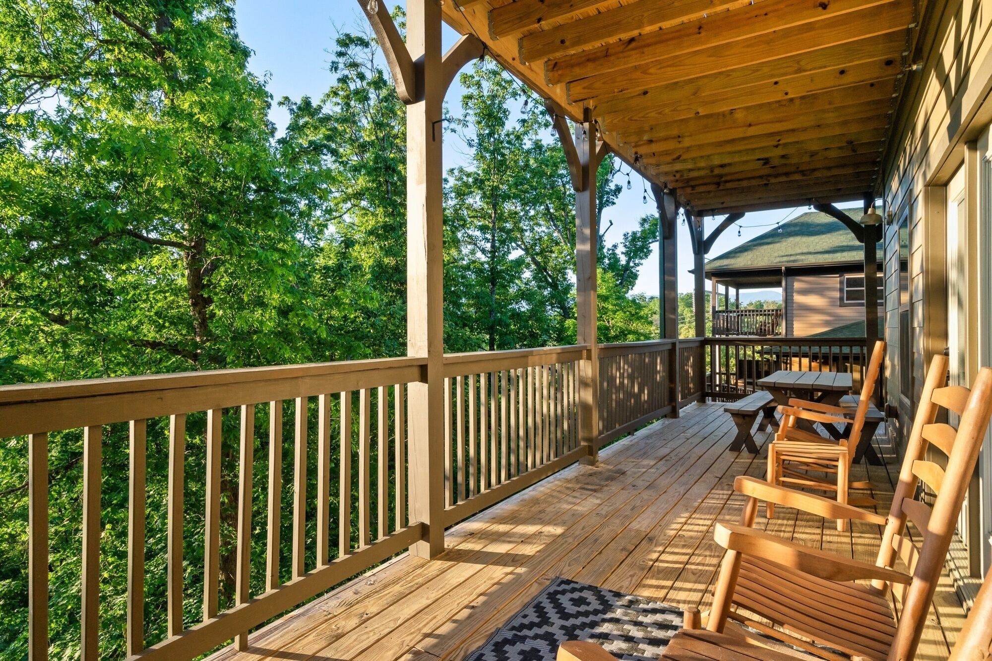 Una terraza de madera con un área cubierta, mobiliario exterior y vistas a una casa al fondo.