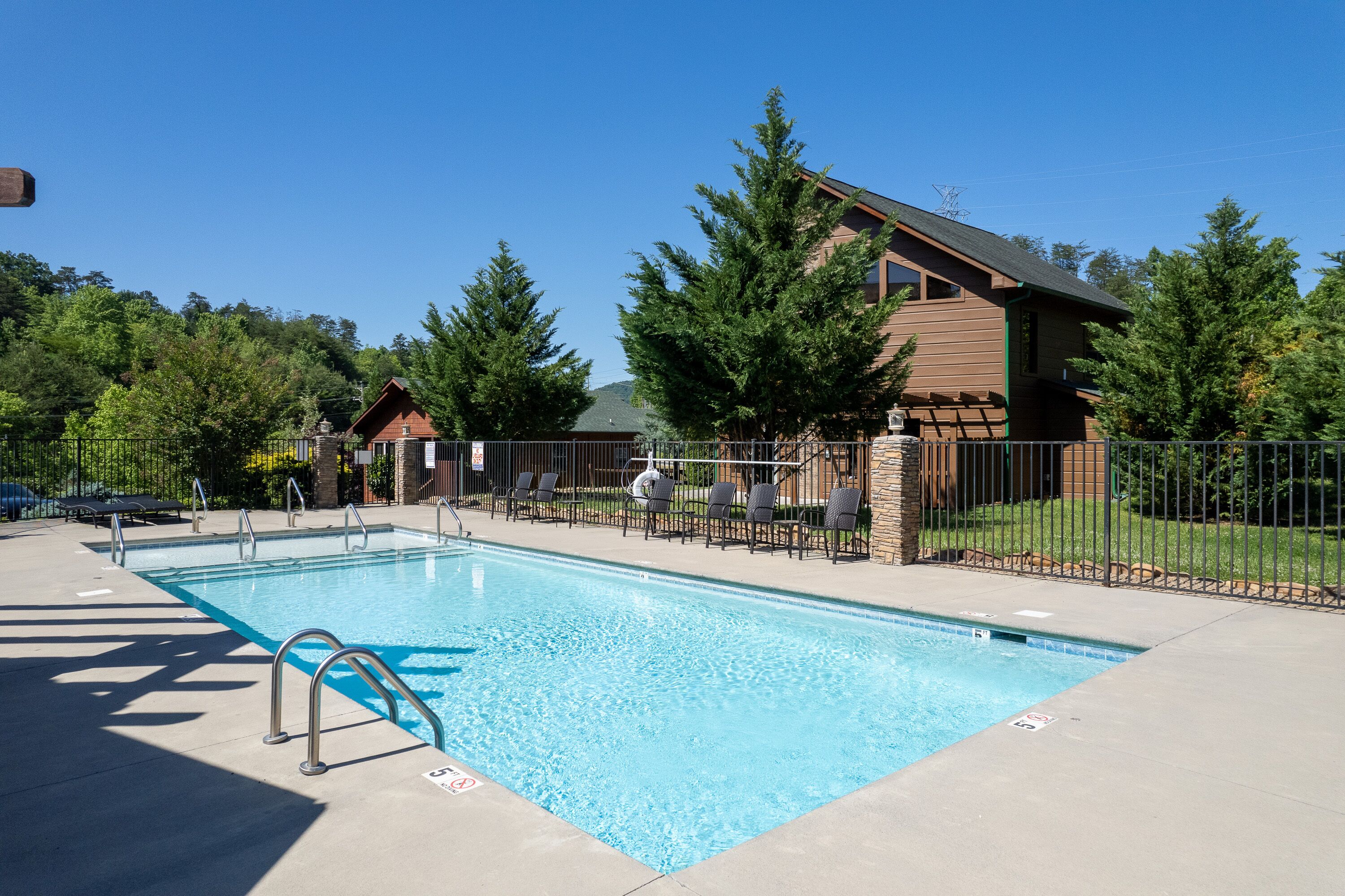 Una piscina rectangular con una escalera metálica, rodeada por un patio de concreto. Se ven sillones de descanso y un edificio de madera con techo inclinado al fondo.
