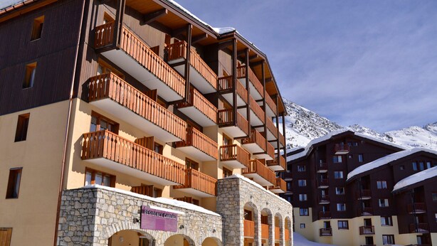 A multi-story hotel with wooden balconies and stone arches, set against a snowy mountain backdrop.