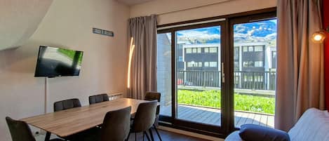 A modern room with a wooden dining table, four chairs, a flat-screen TV, a sofa, and a sliding glass door leading to a balcony with a view of buildings and greenery.