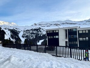 Ein modernes Gebäude mit flachem Dach und großen Fenstern, umgeben von schneebedeckten Bergen und blauem Himmel.
