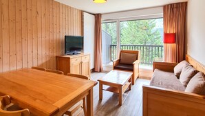 Ein holzmöbliertes Zimmer mit Esstisch, einer Couch, einem Fernseher auf einem Schrank und einem Balkon mit Vorhängen.