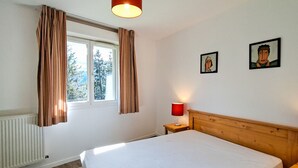Ein Schlafzimmer mit einem Bett, einem Nachttisch, einer Lampe, einem Fenster mit Vorhängen und zwei gerahmten Bildern an der Wand.