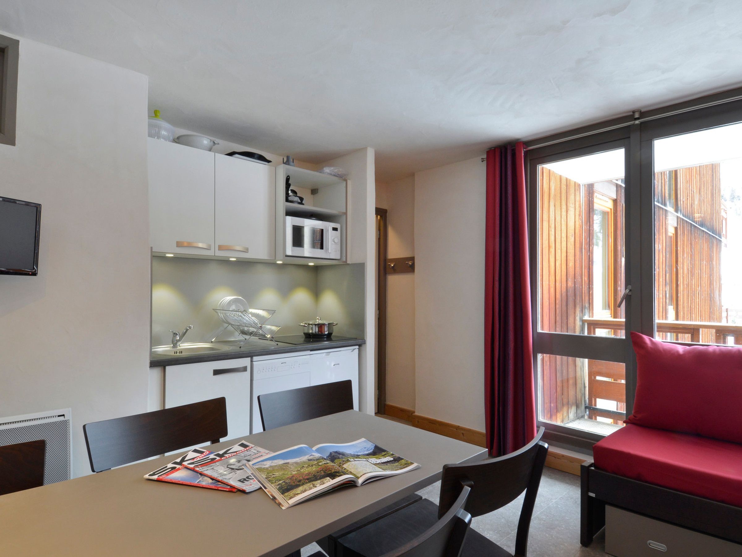 A compact kitchen with white cabinets, a microwave, and a sink. There is a dining table with magazines and a red sofa with a window view.