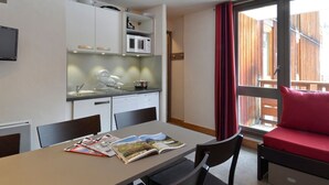 Een compacte keuken met witte kastjes, een magnetron en een spoelbak. Er staat een eettafel met tijdschriften en een rode, beklede zitting.