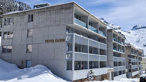 Un immeuble à plusieurs étages, sur lequel on peut lire l’inscription « PETITE OURSE », entouré de neige et de montagnes.