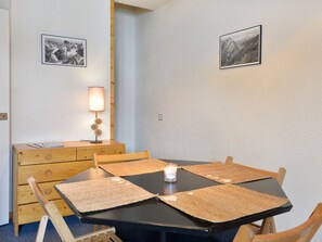 Et spiseområde med et sort bord, fire træstole og to servietter. Et stearinlys står på bordet, og der er et træskab med skuffer til venstre. To billeder hænger på væggene.