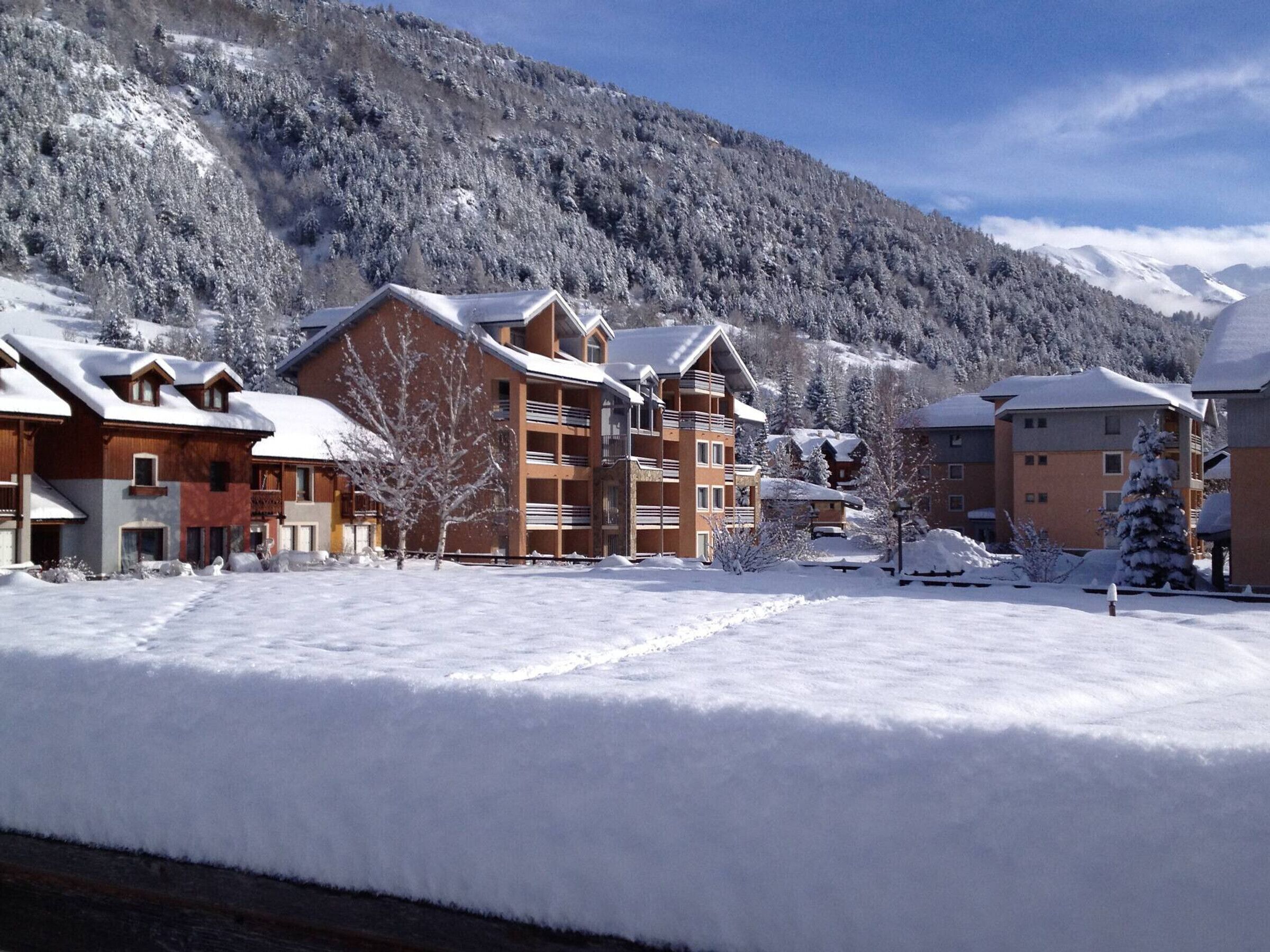 A snow-covered resort with multiple buildings, including a large lodge with balconies and a smaller building with a sloped roof.