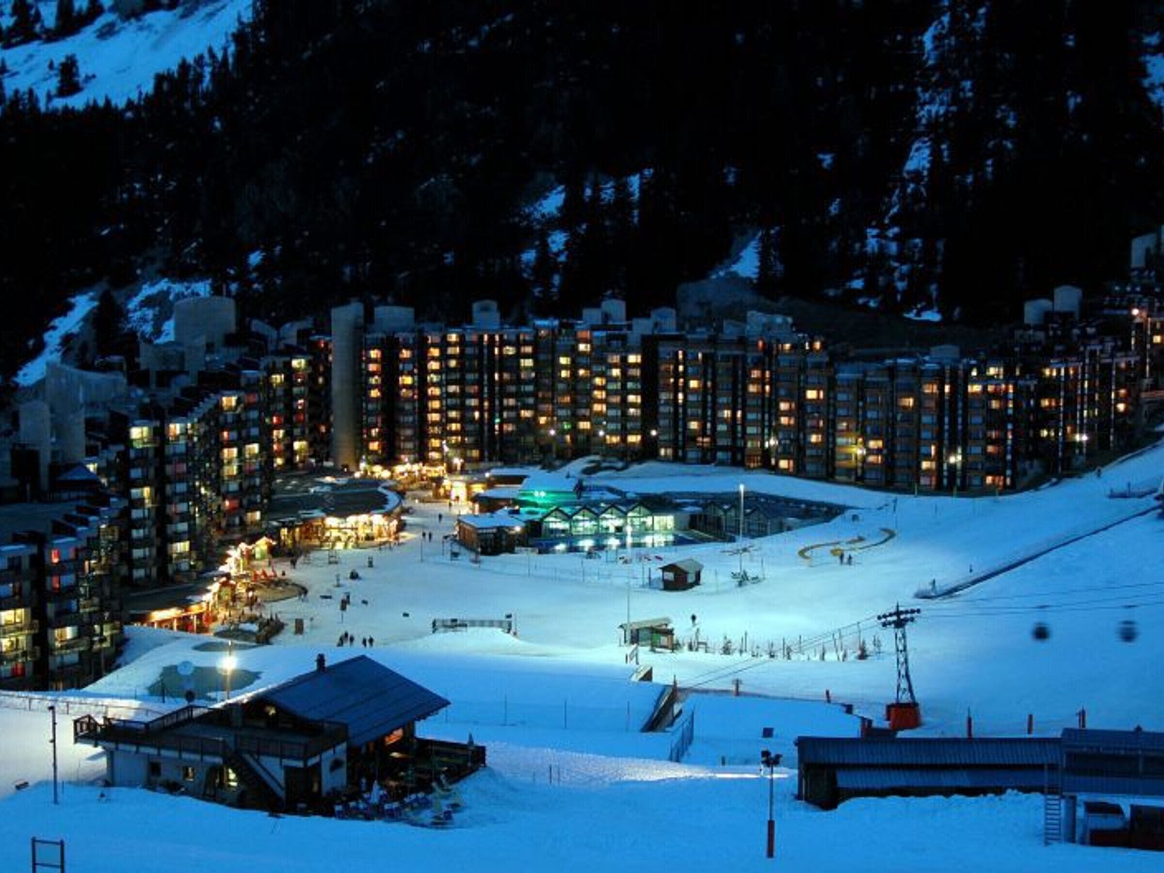Une station de ski enneigée, illuminée la nuit, avec des bâtiments éclairés et un télésiège.