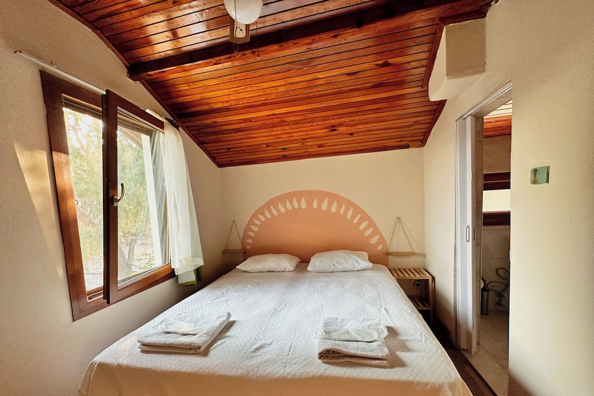 Un dormitorio con techo de madera, una cama con ropa blanca, dos almohadas y una mesita de noche con una lámpara.