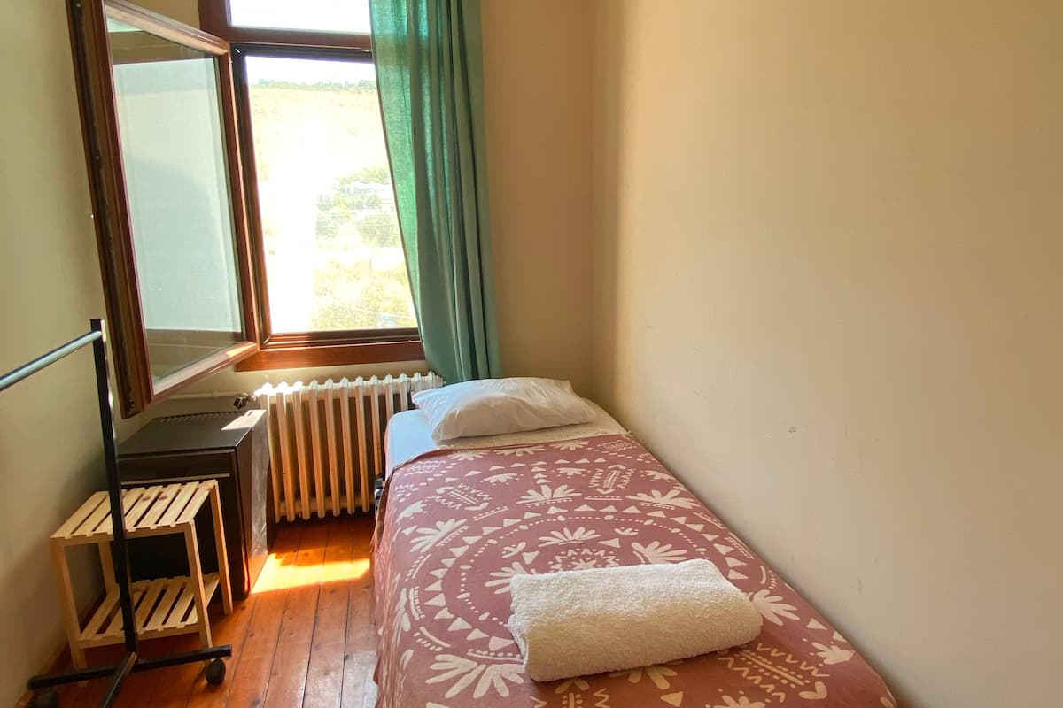 Un dormitorio con una cama, ventilador de techo, radiador, ventana con cortinas y una mesita.
