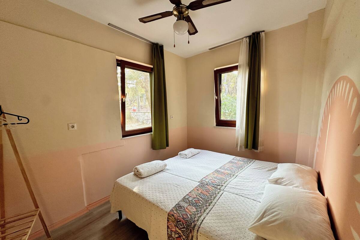 Un dormitorio con una cama, un ventilador de techo, dos ventanas con cortinas y suelo de madera.