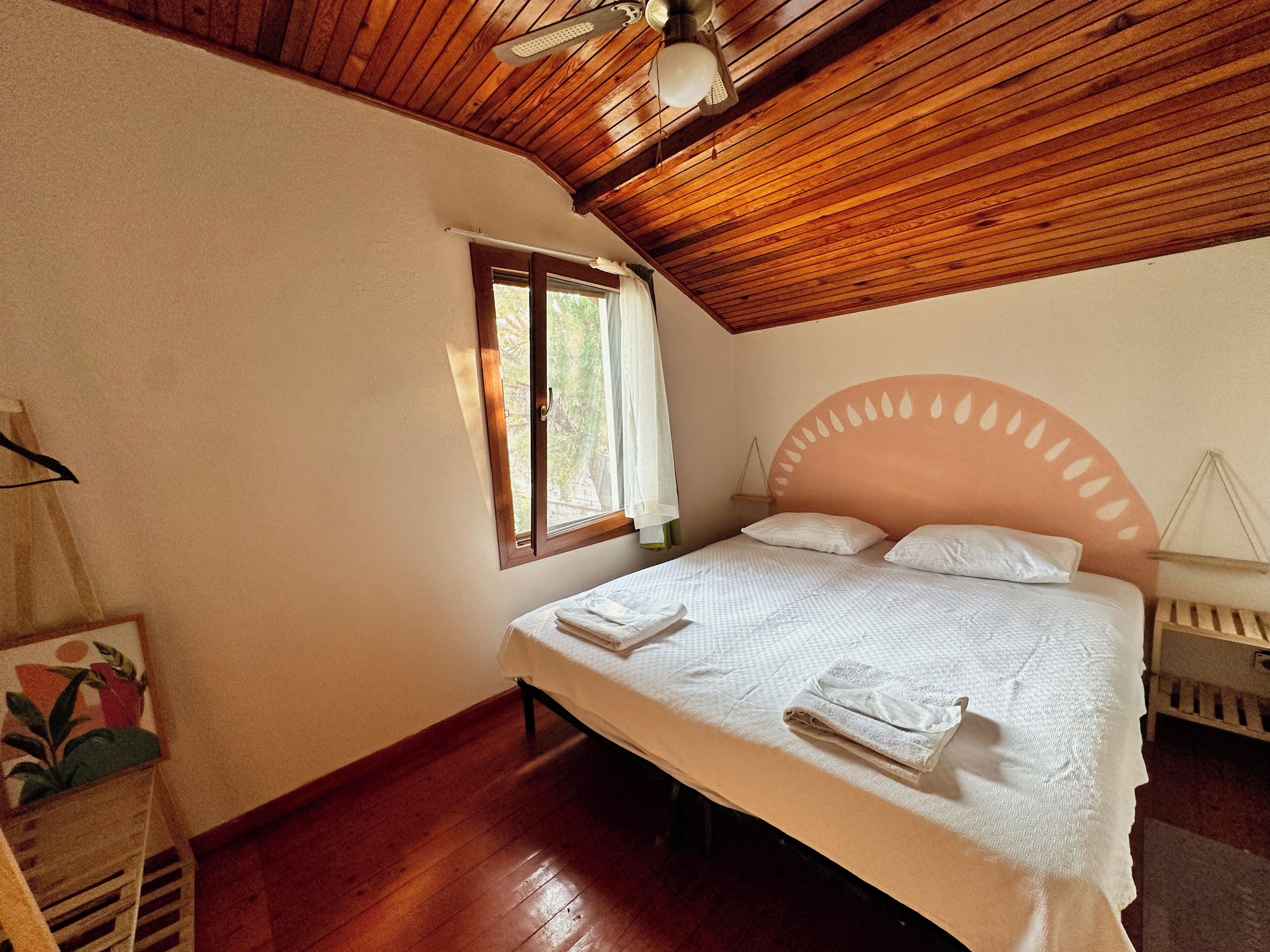Un dormitorio con techo de madera, una cama con ropa de cama blanca, una ventana con cortinas y una mesita de noche pequeña.
