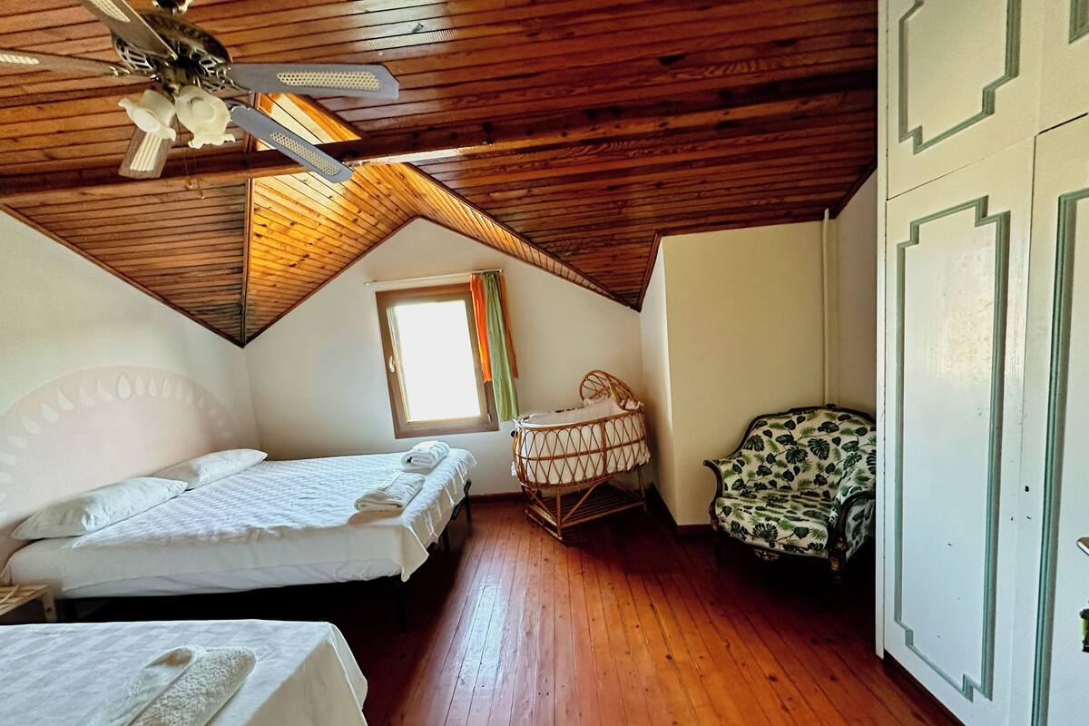 Una habitación con techo inclinado, piso de madera, una cama con sábanas blancas, una cuna pequeña, una silla con estampado de hojas y una ventana con cortinas.