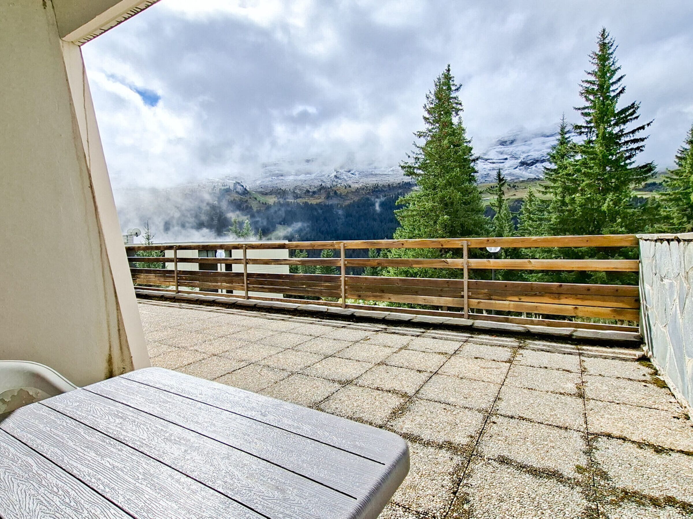 A balcony with a wooden bench and a table, overlooking a mountainous landscape with evergreen trees.