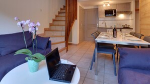 A modern living room with a sofa, a dining table set for two, a laptop, and a staircase leading to a kitchen area. - Comfortable semi-detached chalet 8 pers · Shuttle to slopes · Free WIFI · Terrace (La Salle-les-Alpes)