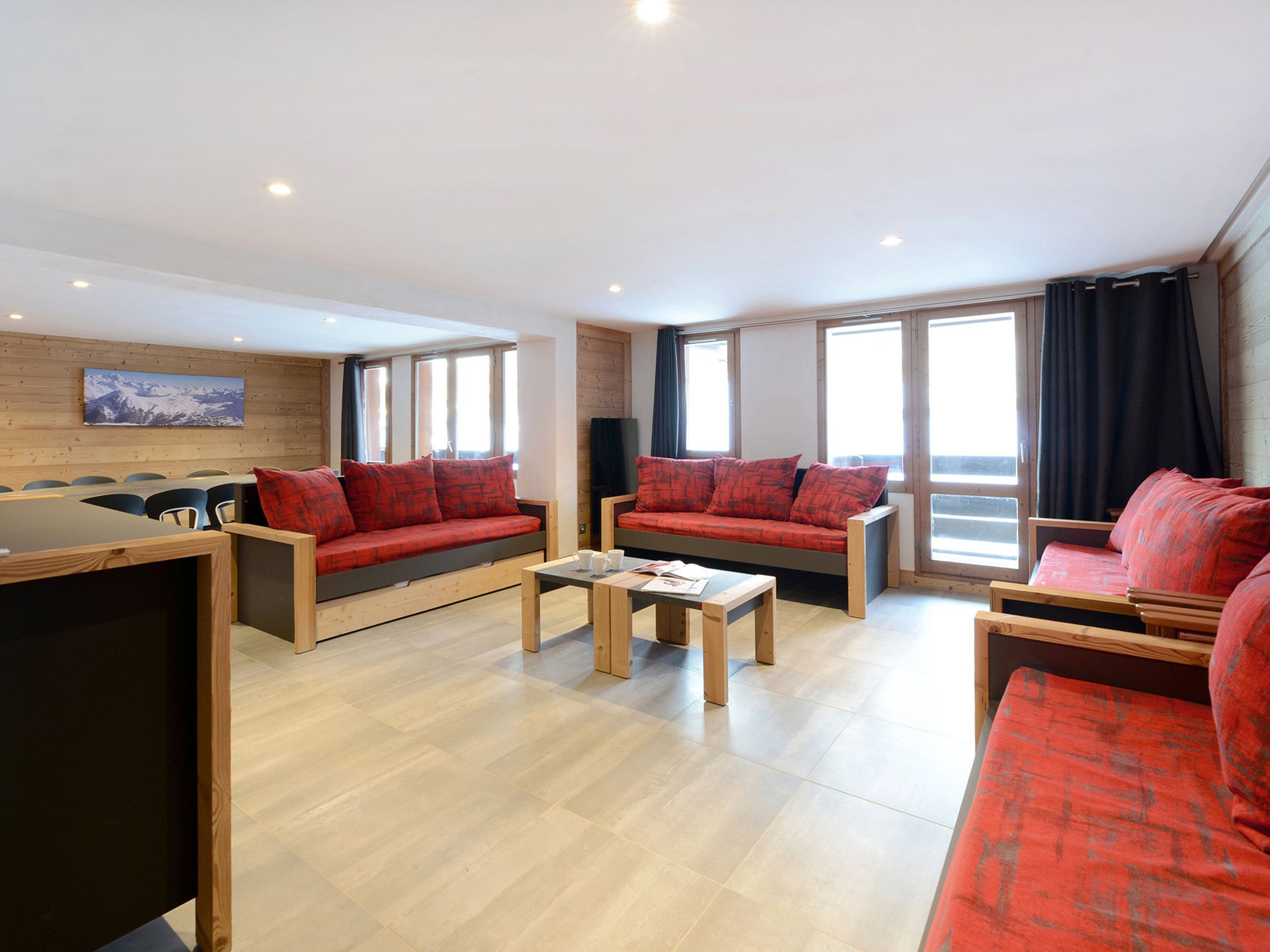 A modern living room with wooden flooring, red sofas, a coffee table, and large windows.
