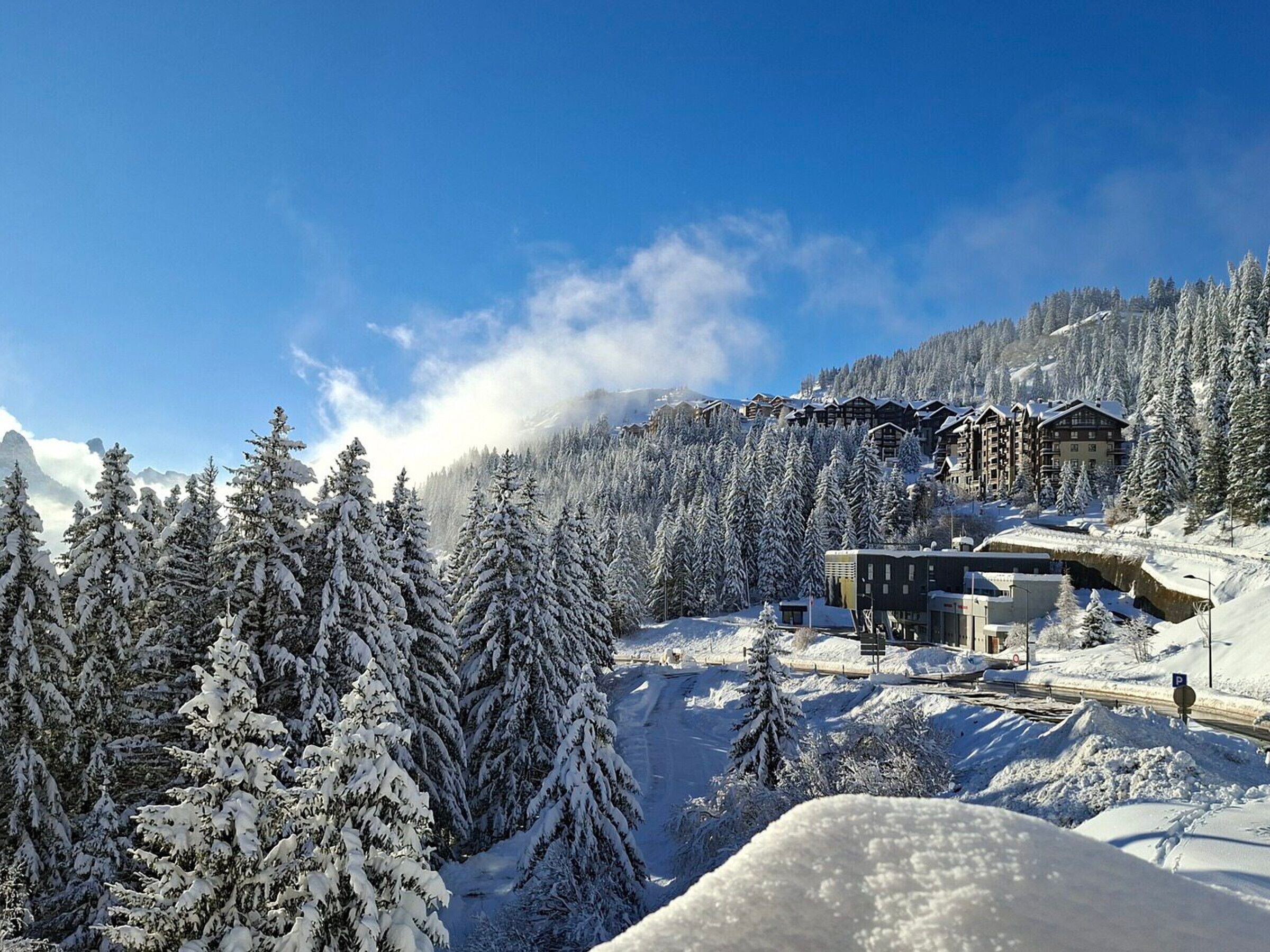 Une station de montagne enneigée, avec des bâtiments d’hébergement, un télésiège et de nombreux conifères.