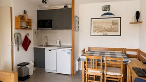 Eine kompakte Küche mit Geschirrspüler, Mikrowelle und Spüle. Es gibt einen Essbereich mit Tisch und Stühlen sowie einen kleinen Holzschrank.
