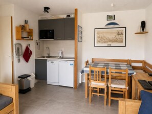 A compact kitchen with a dishwasher, microwave, and sink. There is a dining area with a table and chairs, and a small wooden cabinet.