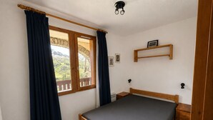 Ein Schlafzimmer mit einem Bett, einem Holzkopfteil, einem Fenster mit Vorhängen, einem Nachttisch, einem an der Wand montierten Regal und einem Bilderrahmen.