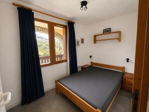 A bedroom with a bed, wooden headboard, a window with curtains, a bedside table, a wall-mounted shelf, and a picture frame.