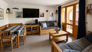 Ein gemütliches Wohnzimmer mit einem Holzesstisch, einer Couch, einem Fernseher und einem großen Fenster mit Vorhängen.