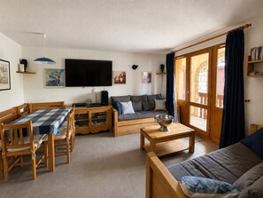 A cozy living room with a wooden dining table, a sofa, a TV, and a large window with curtains.