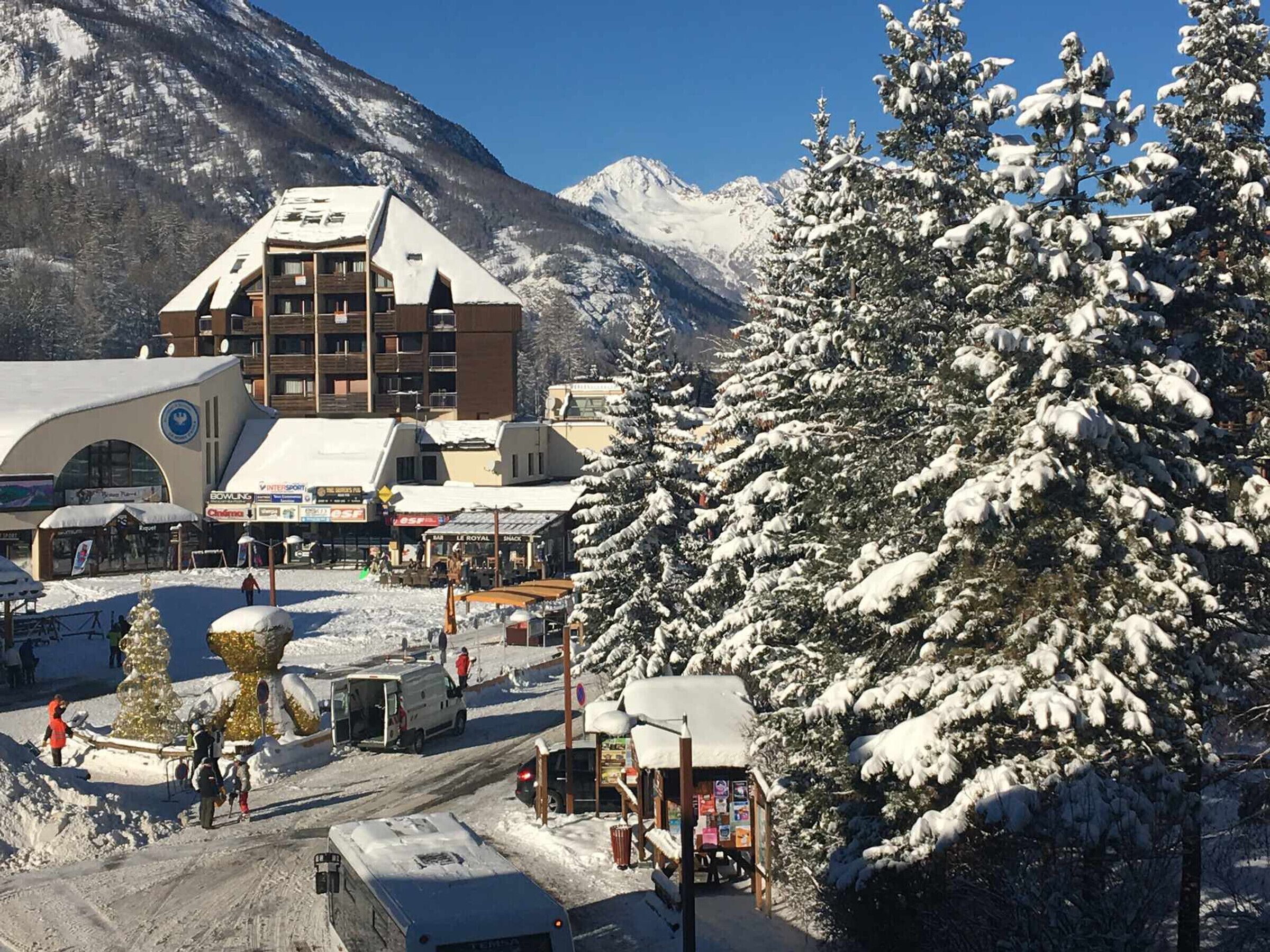 Un pueblo nevado al pie de una montaña, con un edificio destacado que cuenta con un telesilla, rodeado de árboles cubiertos de nieve y un cielo azul despejado.