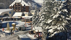 Une ville de montagne enneigée, dominée par un bâtiment imposant qui abrite un télésiège, entourée d’arbres chargés de neige et surplombée par un ciel bleu clair.