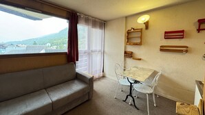 Une pièce comprenant un canapé, une table avec des chaises, une fenêtre avec des rideaux et des étagères fixées au mur.