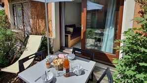 An outdoor dining area with a table set for two, a wicker basket with bread, a bottle of juice, and a glass of water. - Bright triplex chalet, shuttle to slopes, Wifi, pets allowed (La Salle-les-Alpes)