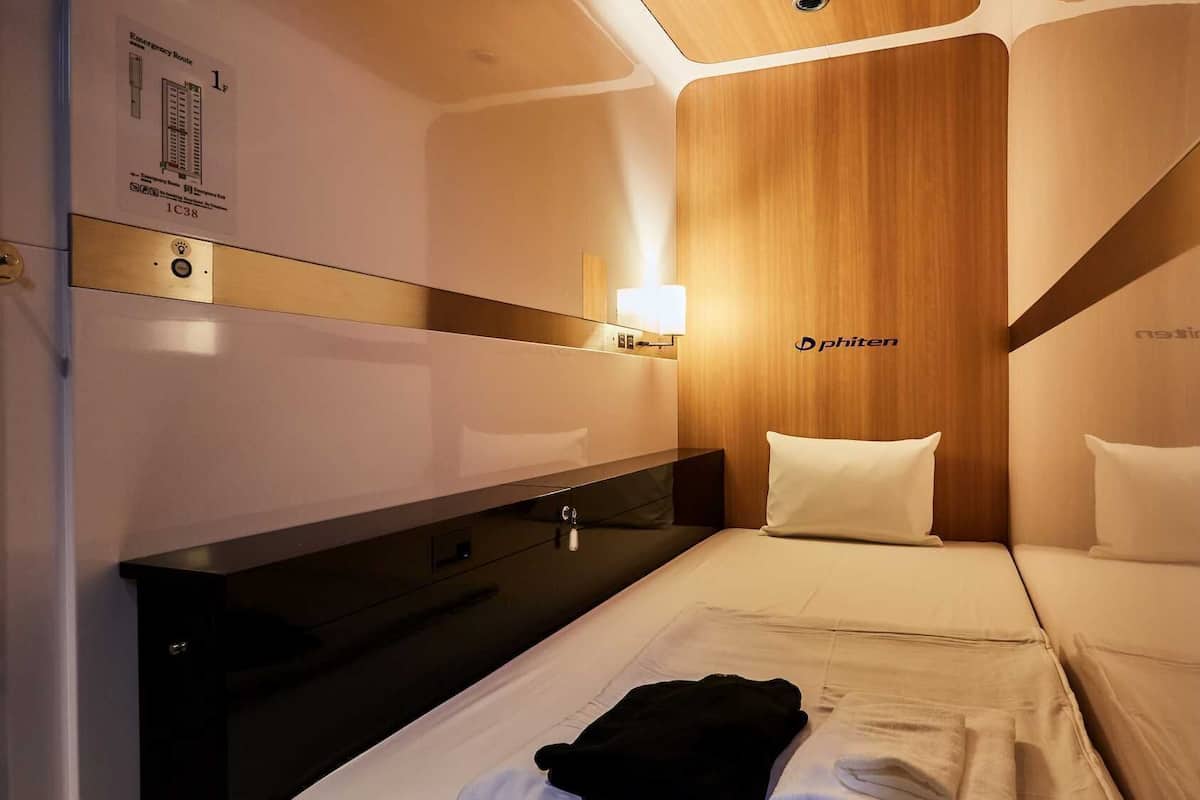 Une chambre d’hôtel de taille réduite, comprenant un lit, une table de chevet, un miroir, une lampe fixée au mur et un mur recouvert de panneaux en bois.