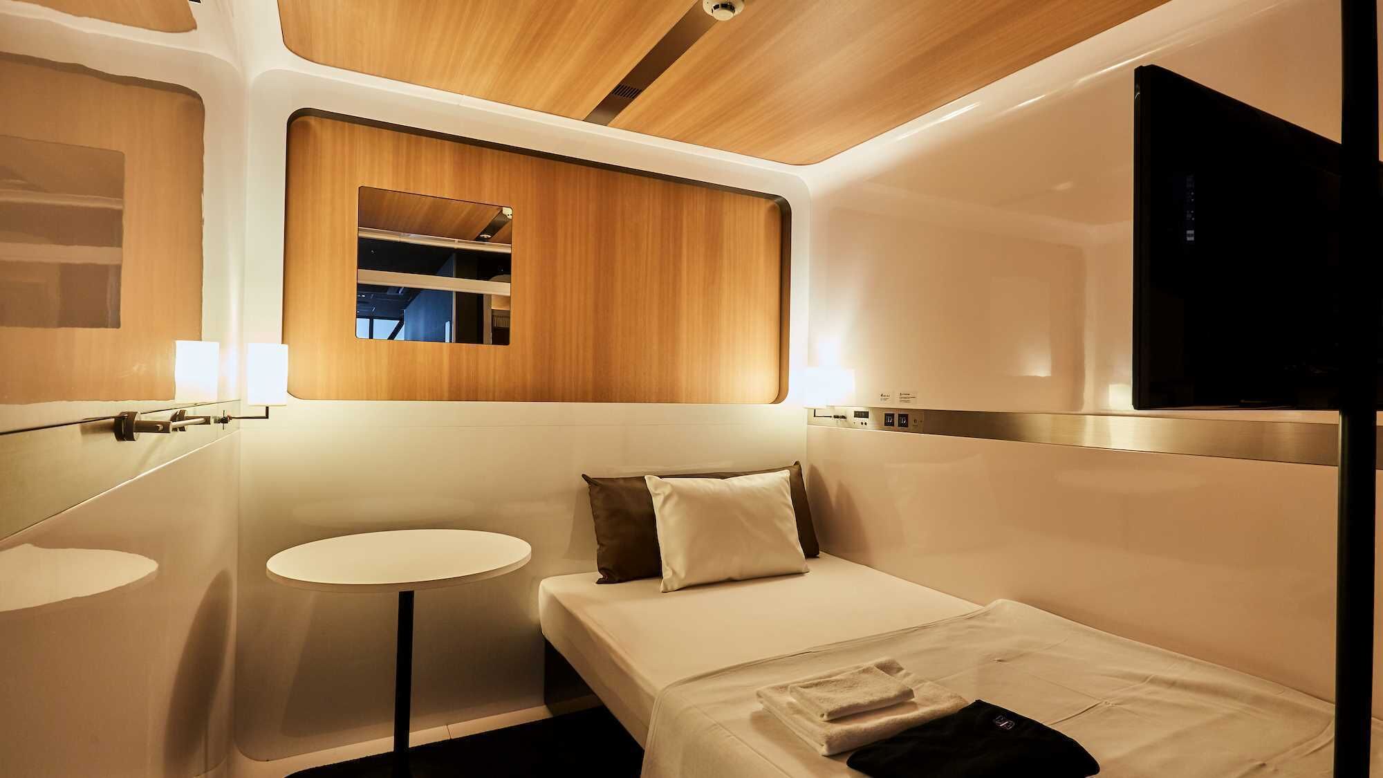 A compact, modern hotel room with a bed, bedside table, mirror, and a small round table.