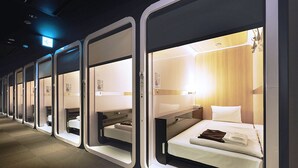 A row of private sleeping pods with individual beds, each pod has a small table and a chair, and a window. - First Cabin Nishi Umeda (Osaka)