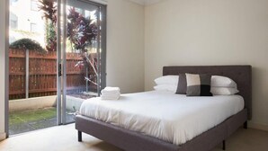 Una camera da letto con un grande letto, una porta scorrevole in vetro che dà su un giardino e vista su una recinzione di legno e piante.