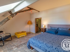 A bedroom with a sloped ceiling, a bed with patterned bedding, a bedside table with a lamp, a yellow bench, and a chair.